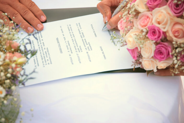 Close-up of a person signing a document with a bouquet of pink roses nearby, hands visible on the page.